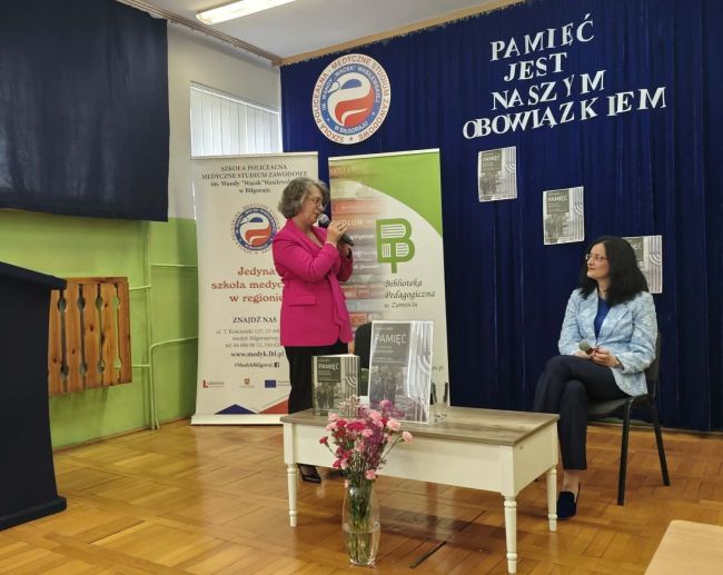 rozmawiające ze sobą kobiety, jedna stoi i mówi do mikrofonu, druga siedzi