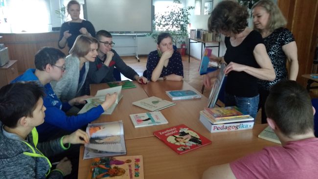Uczestnicy zajęć siedzą przy stoliku i przyglądają się książkom, które prezentuje bibliotekarz prowadzący zajęcia.