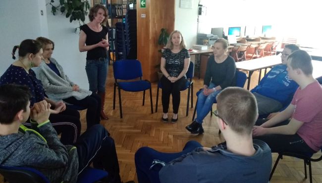 Uczestnicy siedzą w kręgu. Prowadząca na stojąco omawia tematykę zajęć.