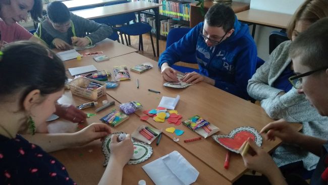 Uczniowie siedzą przy stoliku i wykonują pracę plastyczną – dekorują papierowe serca.