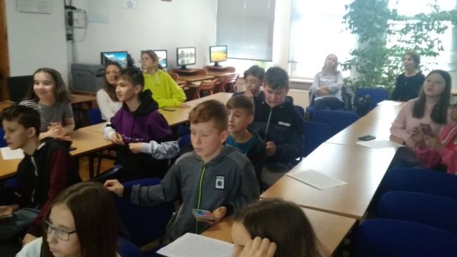 Uczniowie siedzą przy stolikach i rozwiązują quiz z użyciem smartfonów