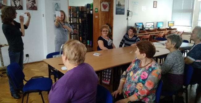 Seniorzy siedzą przy stole. Prowadzące prezentują karteczki z rysunkami.