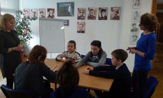 Uczniowie siedzą przy stole. Dwie prowadzące tłumaczą wykonanie zadania. 