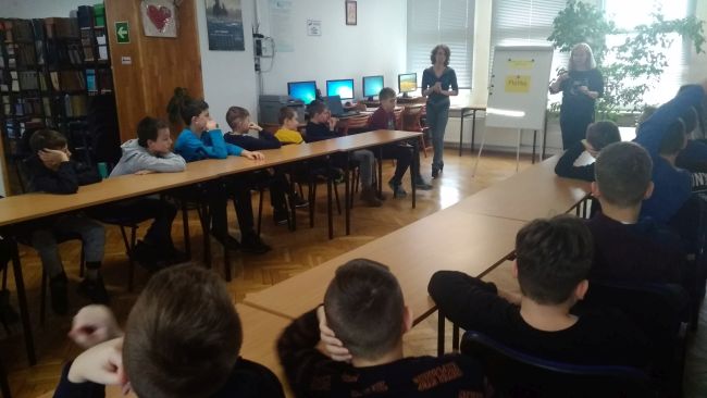 Uczniowie siedzą w ławkach. Nauczyciele bibliotekarze omawiają tematykę zajęć.