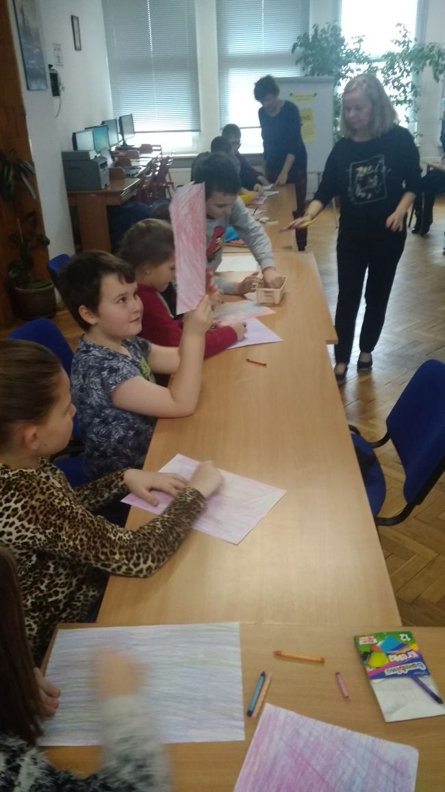 Uczniowie siedzą w ławkach i kolorują kredkami kartki papieru.