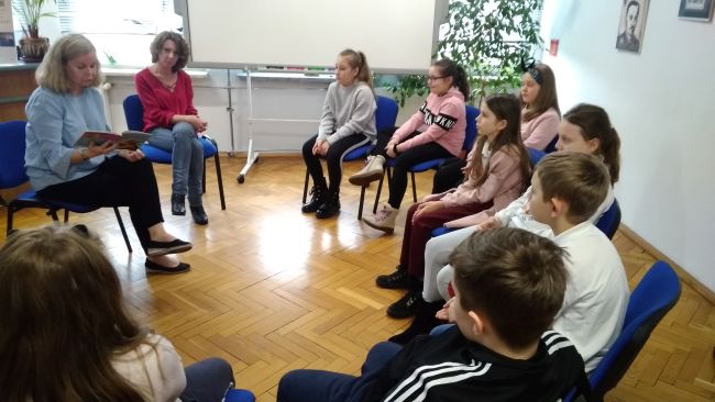 Uczniowie klasy trzeciej siedzą na krzesłach ustawionych w kręgu i słuchają prowadzącej, która czyta książkę pt. 