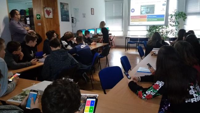 Nauczyciel bibliotekarz prowadzi interaktywny quiz. Uczniowie siedzą w ławkach i udzielają odpowiedzi za pomocą smartfonów.