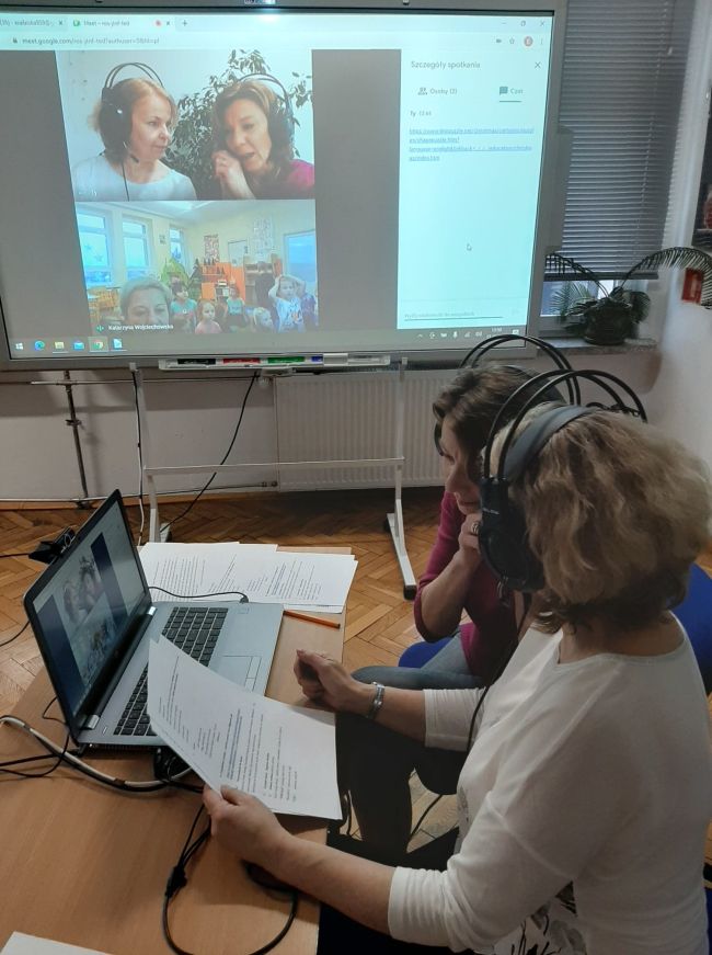 Biblioteka. Przy stoliku pracownicy prowadzący zajęcia on-line. W tle tablica interaktywna z widokiem prowdzących oraz grupy przedszkolaków