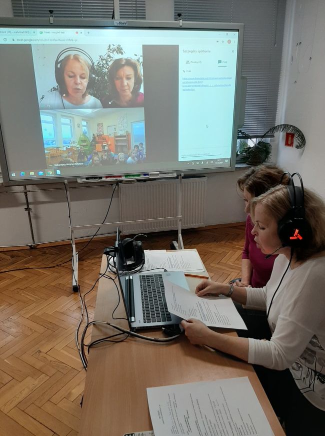 Biblioteka. Przy stoliku pracownicy prowadzący zajęcia on-line. W tle tablica interaktywna z widokiem prowdzących oraz grupy przedszkolaków