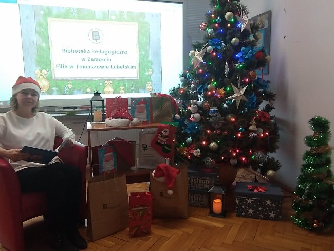W lewym rogu kierownik biblioteki Elżbieta Rafalska czytająca bajkę. W prawym rogu choinka. W centralnej części tablica interaktywna z logo biblioteki oraz prezenty