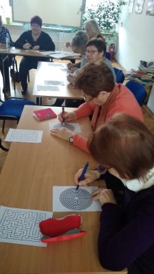 Seniorzy przy stolikach rozwiązują wydrukowany na kartce labirynt.