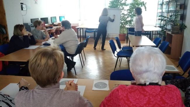 Seniorzy siedzą w ławkach. Prowadząca zapisuje na flipchardzie polecenie do kolejnego zadania.