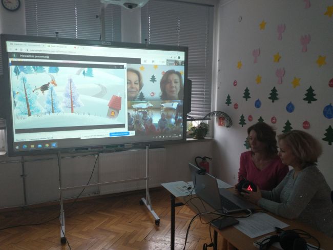 Biblioteka. Przy stoliku prowadzący zajęcia on-line. W tle tablica interaktywna z z widokiem prowadzących oraz grupy przedszkolaków