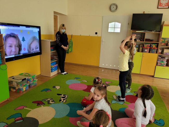 Sala przedszkolna. Grupa dzieci siedzi na dywanie wykonując polecenia prowadzących, widocznych na ekranie telewizora