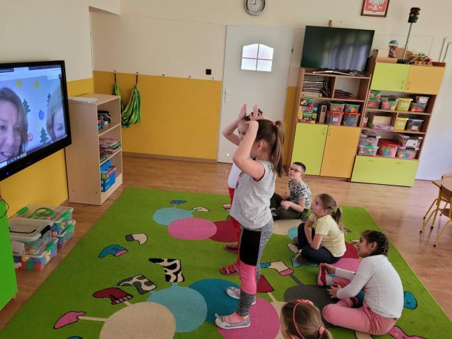 Sala przedszkolna. Grupa dzieci siedzi na dywanie wykonując polecenia prowadzących, widocznych na ekranie telewizora
