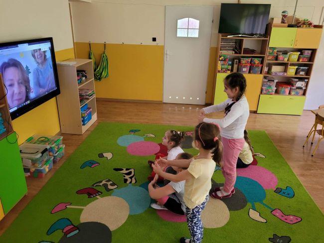 Sala przedszkolna. Grupa dzieci siedzi na dywanie wykonując polecenia prowadzących, widocznych na ekranie telewizora