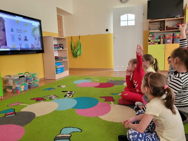 Sala przedszkolna. Grupa dzieci siedzi na dywanie wykonując polecenia prowadzących, widocznych na ekranie telewizora