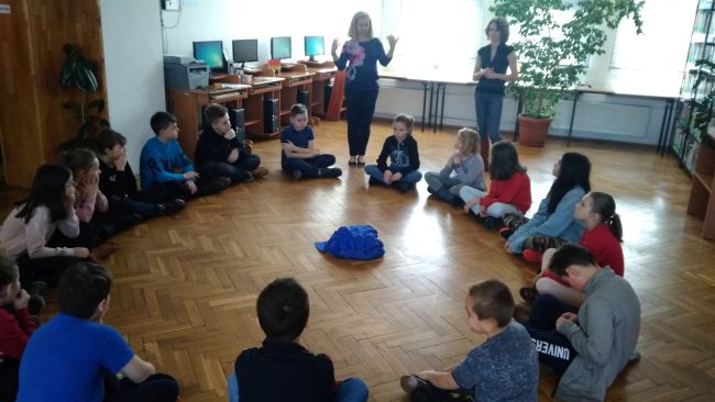 Uczniowie siedzą na podłodze w kręgu. Bibliotekarki prowadzące zajęcia na stojąco witają uczniów i wprowadzają w tematykę zajęć