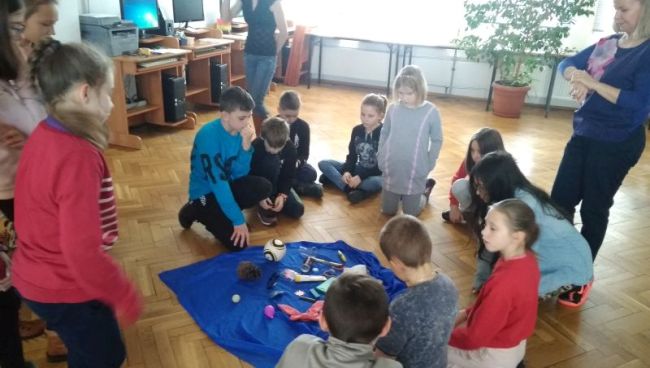 Uczniowie skupieni są wokół chusty rozłożonej na podłodze, na której rozrzucone są różne drobne przedmioty.