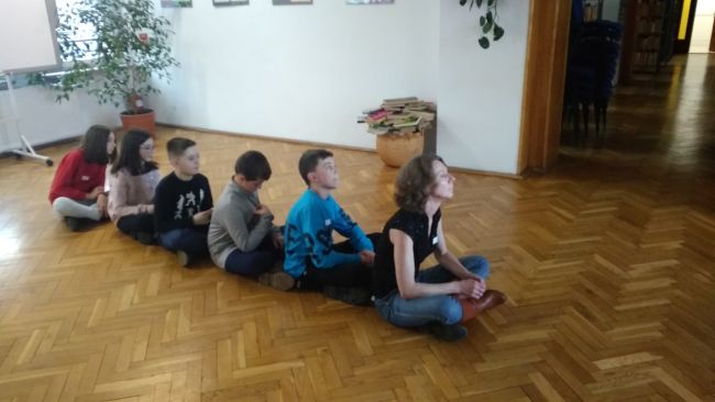 Uczniowie wraz z prowadzącą siędzą po turecku na podłodze, w rzędzie 