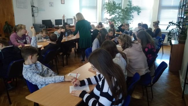 Uczniowie siedzą w ławkach i rozwiązają na kartkach zadania. Nauczyciel bibliotekarz podchodzi do uczniów.