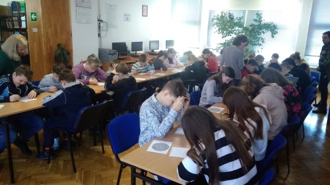 Uczniowie siedzą w ławkach i rozwiązają na kartkach zadania.