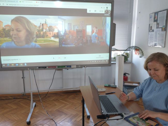 Nauczyciel bibliiotekarz prowadzącego zajęcia on-line. Na tablicy interaktywnej, znajdującej się obok, widać fotografię Wawelu i dzieci wykonujące zadania związane z tematem zajęć.