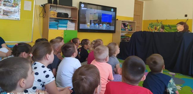 Przedszkolaki siedzą na dywanie i oglądają teatr kukiełkowy prezentowany przez nauczycieli bibliotekarzy