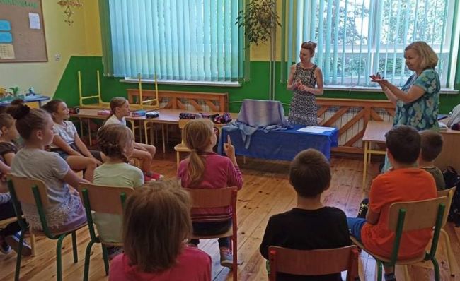 Uczniowie siedzą na krzesełkach w półkolu, w klasie szkolnej. Przed dziećmi nauczyciele bibliotekarze prowadzący zajęcia.