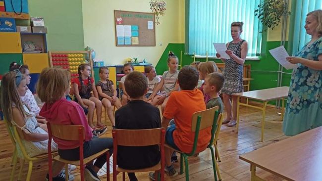Dzieci siedzą na krzesełkach w kręgu i odgadują zagadki czytane przez nauczycieli.
