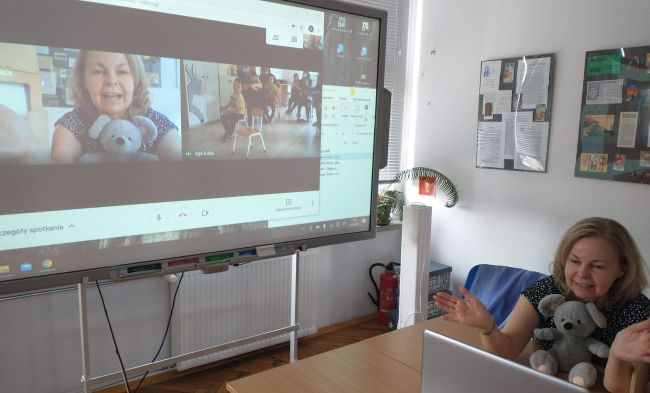 Biblioteka. Przy stoliku prowadząca zajęcia on-line. W tle tablica interaktywna z widokiem grupy przedszkolaków