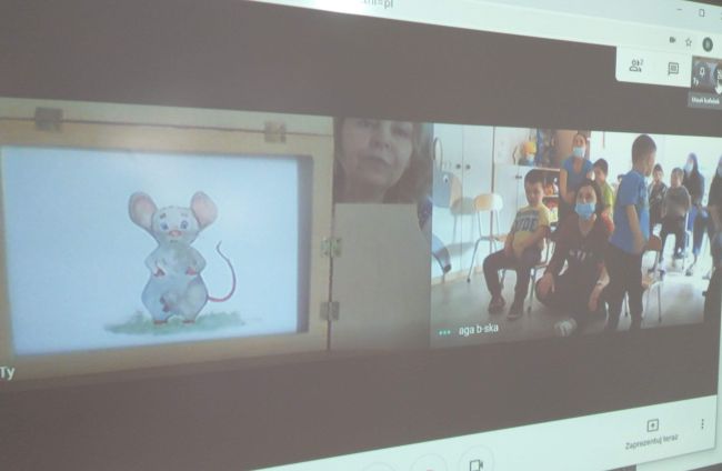 Biblioteka. Przy stoliku prowadząca zajęcia on-line z maskotką - pluszową myszką .W tle tablica interaktywna z widokiem teatru Kamishibai