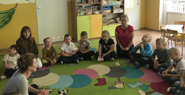 Dzieci siedzą na dywanie, słuchają prowadzącej tłumaczącej zadanie. Przygotowują się do wykonywania ćwiczeń i gier na tablicy interaktywnej, związanych ztematem zajęć.