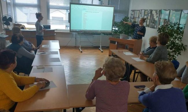Prowadzące omawiają zadanie wyświetlone na tablicy interaktywnej. Seniorzy siedzą w ławkach, czytają ćwiczenie.