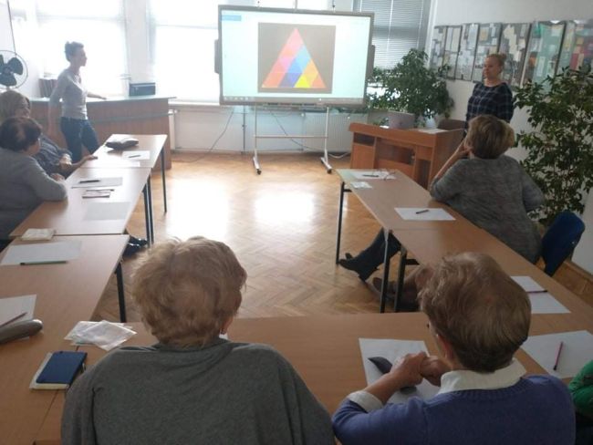 Seniorzy siedzą w ławkach. Rozwiązują zadanie matematyczne. Na tablicy interaktywnej wyświetlony jest duży trójkąt złożony z mniejszych trójkątów. Prowadzące omawiają zadanie.