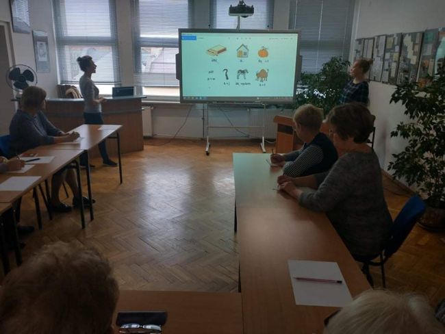 Seniorzy siedzą w ławkach, rozwiązują rebus, wyświetlony na tablicy interaktywnej. 