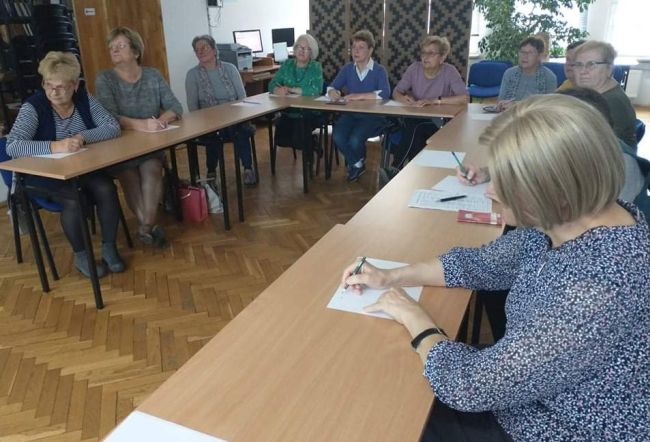 Słuchacze Uniwersytetu Trzeciego Wieku siedzą w ławkach, rozwiązują zadania na kartkach papieru.