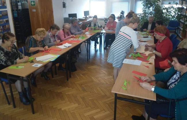 Seniorzy siedzą przy stolikach, wykonują gwiazdy z papieru metodą origami.
