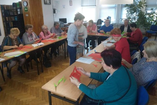 Seniorzy siedzą przy stolikach, wykonują gwiazdy z papieru metodą origami.
