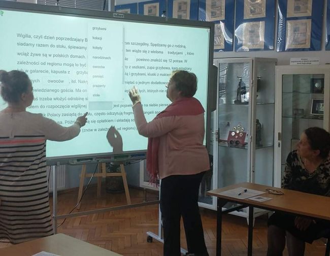 Jedna osoba pisze na tablicy interaktywnej. Prowadząca zajęcia wskazuje polecenia do zadania.