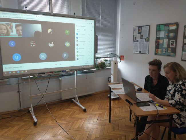 Biblioteka. Przy stoliku prowadzące zajęcia on-line. W tle tablica interaktywna z uczestnikami spotkania