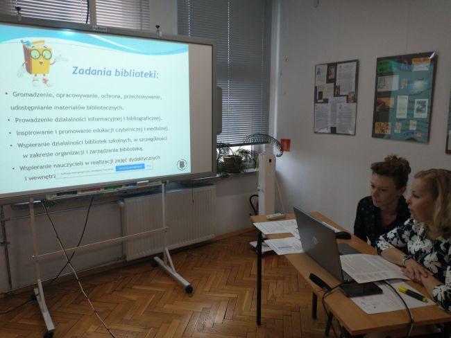 Biblioteka. Przy stoliku prowadzące zajęcia on-line oraz tablica interaktywna z  prezentacją multimedialną
