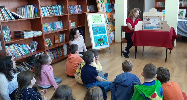Uczniowie siedzą na podłodze w sali biblioteki szkolnej. Prowadząca siedzi przy stoliku i czyta opowieść metodą Kamishibai.