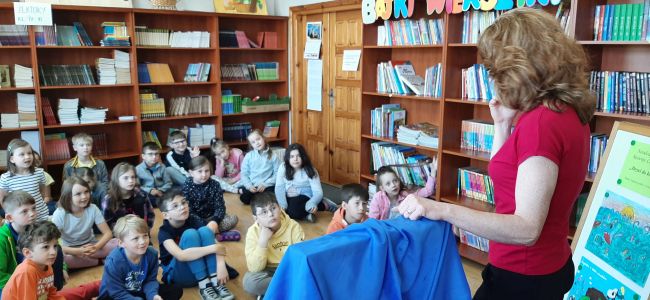 Uczniowie siedzą na podłodze w sali biblioteki szkolnej. Prowadząca stoi i prezentuje skrzynkę Butai.