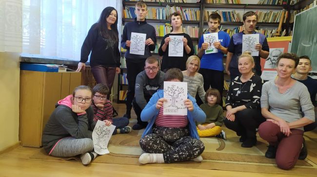 Uczniowie po zajęciach, razem z dwoma nauczycielami i dwiema bibliotekarkami z Biblioteki Pedagogicznej w Tomaszowie Lubelskim w pozują do wspólnego zdjęcia. W rękach trzymają swoje prace plastyczne – jesienne drzewa z kolorowymi liśćmi.
