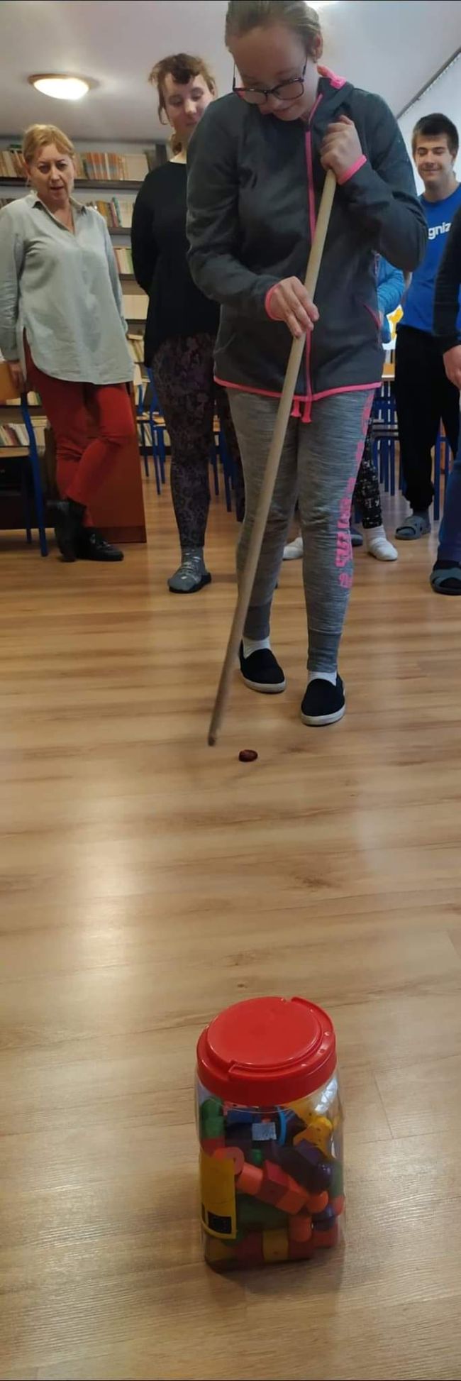 Uczennica pokonuje tor przeszkód. W ręku trzyma kij i przesuwa nim kasztan po podłodze. W tle, za jej plecami  widać innych uczniów.