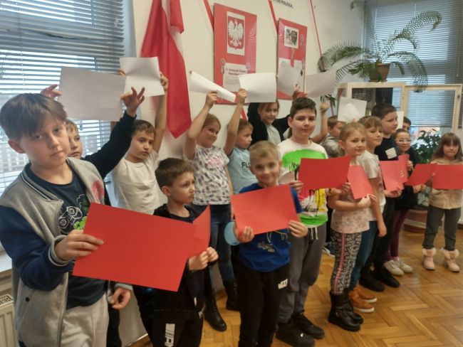 Na zdjęciu uczniowie stoją w dwuszeregu z biało-czerwonymi kartkami imitującymi flagę. W tle wystawa przygotowana z okazji 11 Listopada 
