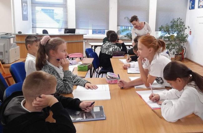 Uczniowie siedzą przy stolikach prowadzące, w pozycji stojącej omawiają temat zajęć