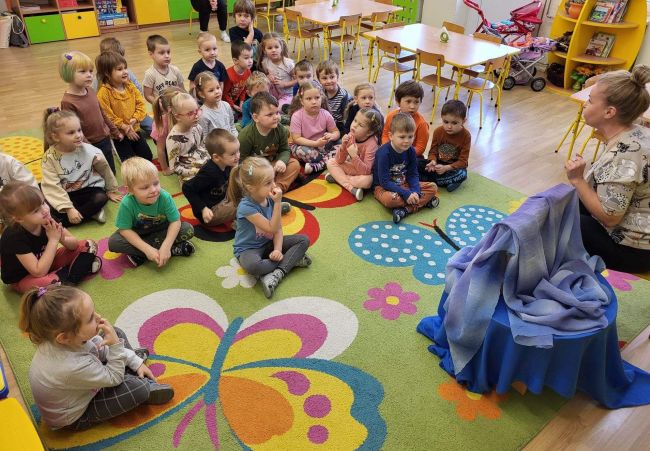 Sala przedszkolna. Dzieci siedzą na dywanie w oczekiwaniu na bajkę. Przed nimi na stoliku ukryty teatrzyk Kamishibai. Obok nauczyciel bibliotekarz prowadzący zajęcia.