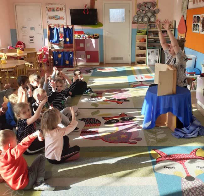 Sala przedszkolna. Dzieci siedzą na dywanie z rękami uniesionymi do góry. Przed nimi na stoliku teatrzyk Kamishibai. Obok nauczyciel bibliotekarz prowadzący zajęcia.